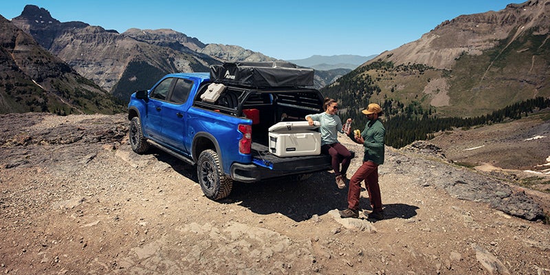 A 2025 Chevrolet Silverado 1500 parked on rocky terrain with the tailgate down, one person sitting on the tailgate opening a cooler, while a gentleman is standing nearby.